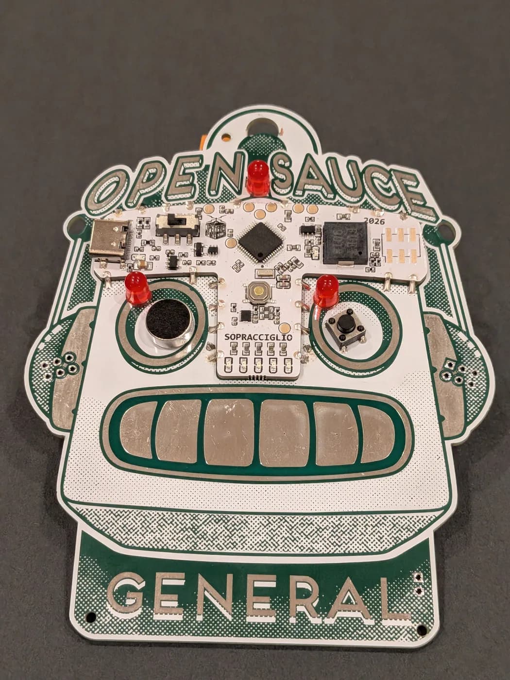A fully assembled Open Sauce 2026 SauceBot badge with microphone, button, and red LEDs installed